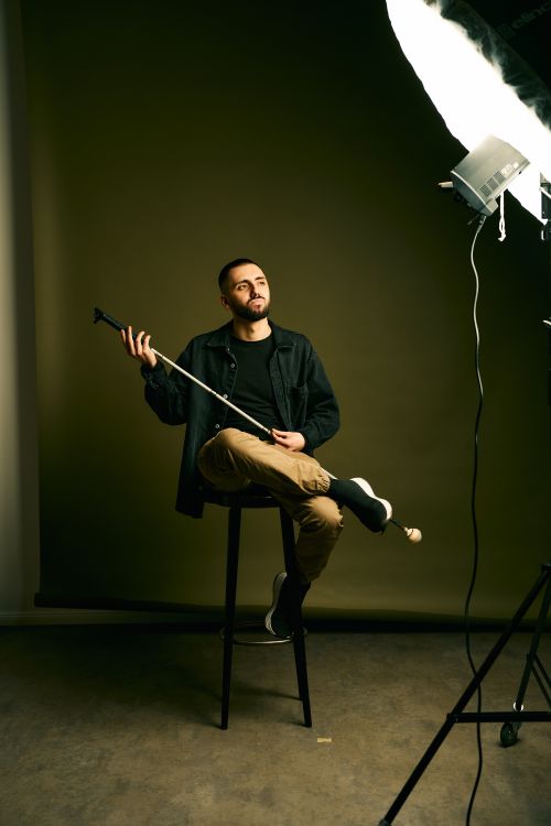 Timur Turga sitzt mit einem coolen Blick auf einem Barhocker in einem Raum der nur von einer hellen Fotografen-Lampe beleuchtet wird und hält seinen Stock wie beim spielen einer Gitarre